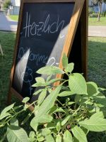 Schief stehende Tafel mit handgeschriebener Begrüßung 'Herzlich willkommen' und grünen Blättern im Vordergrund