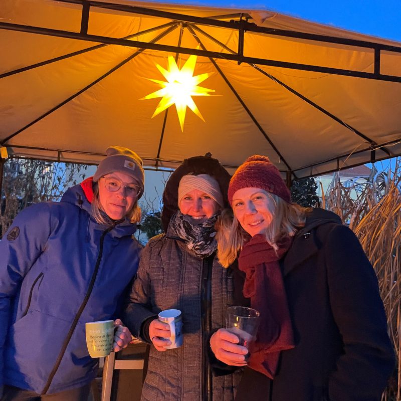 Drei Frauen in Winterkleidung unter einem Pavillon mit leuchtendem Stern, die Tassen und ein Glas halten