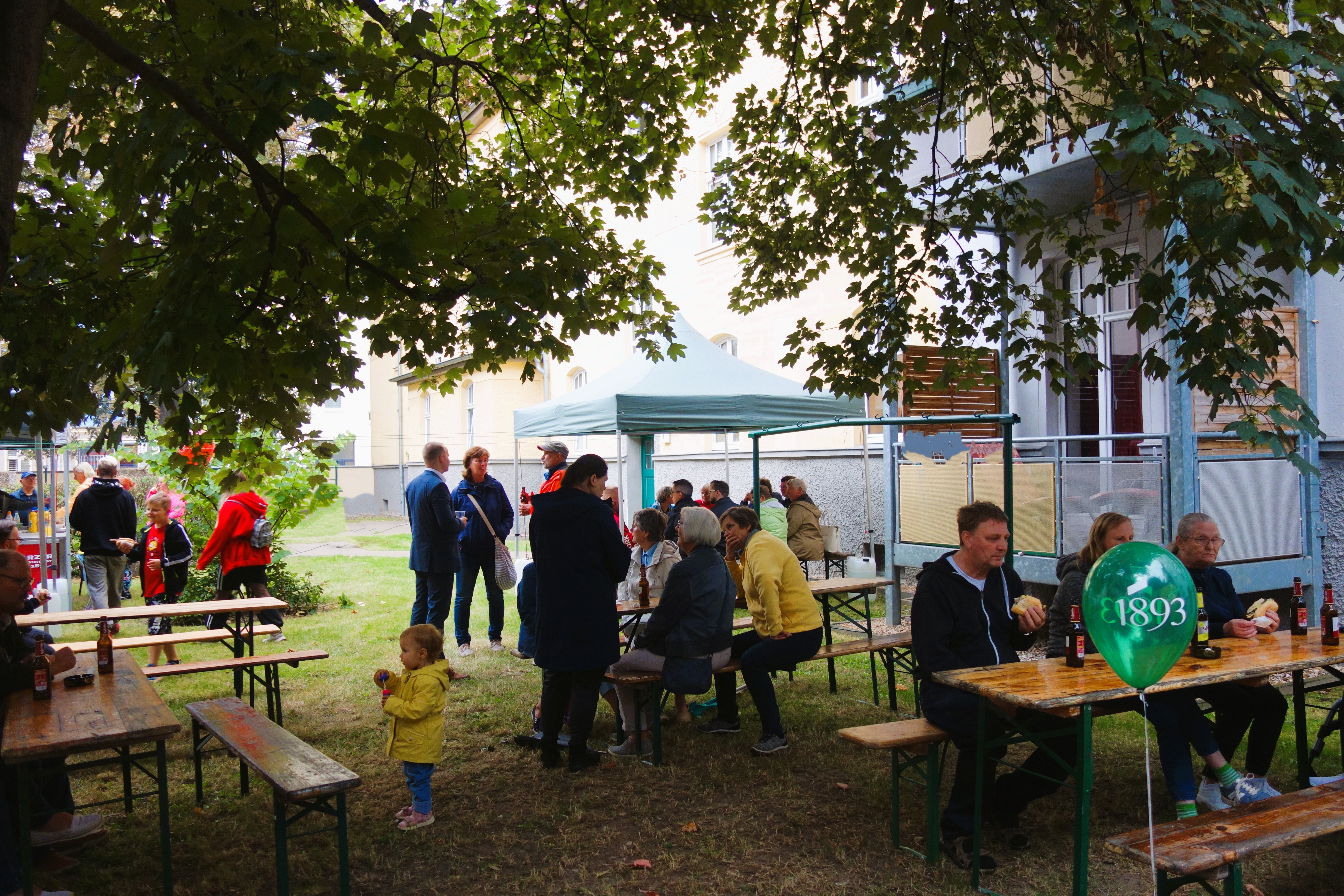 Mehrere Menschen sitzen an Bierbänken unter Bäumen. Im Hintergrund ist ein Wohngebäude.