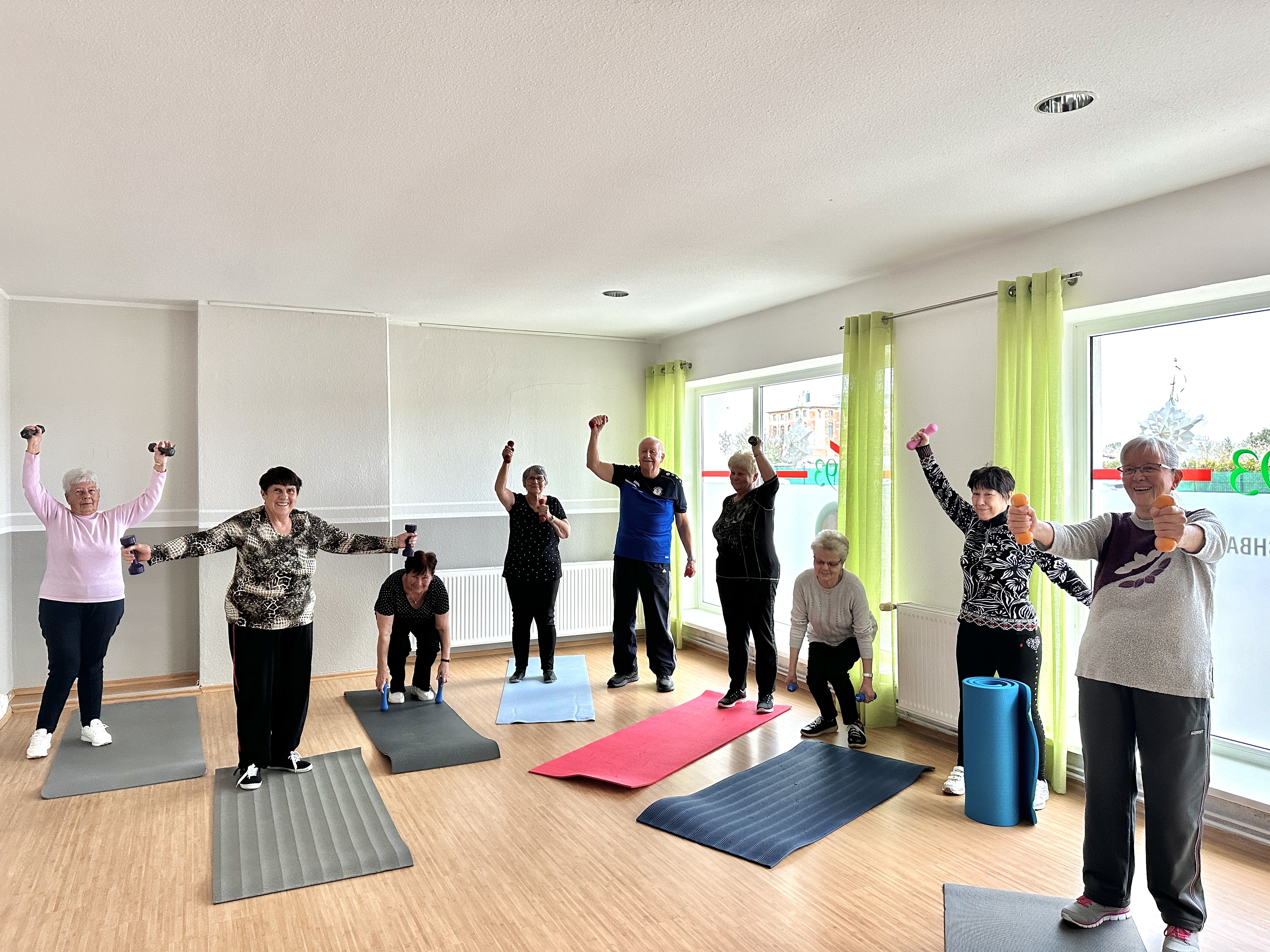 Mehrere Senioren halten Hanteln in den Händen und stehen damit auf Gymnastikmatten in einem hellen Raum. Alle lachen.