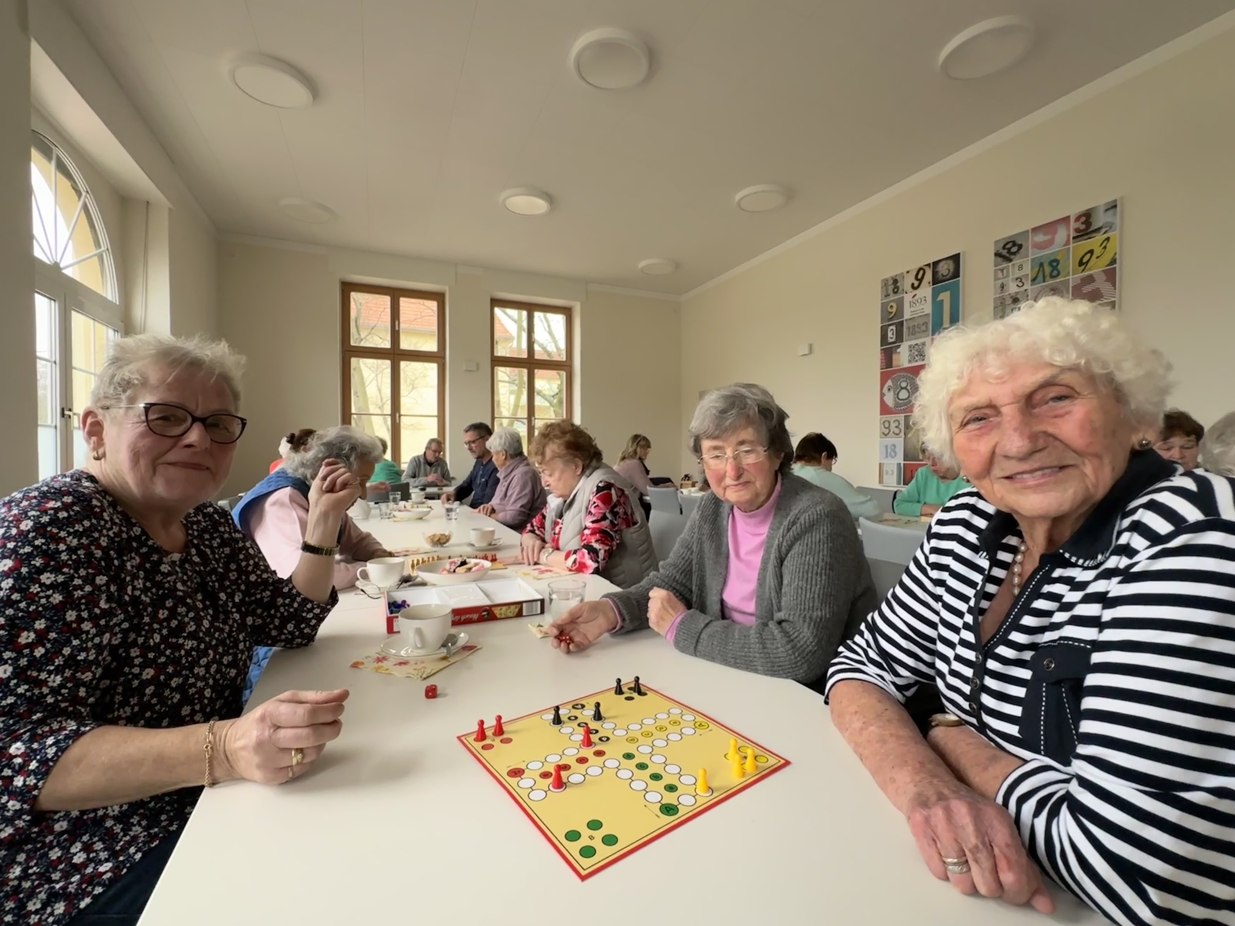 Mehrere ältere Frauen sitzen an einem Tisch und spielen Ludo. Im Hintergrund sind weitere spielende Menschen zu sehen.