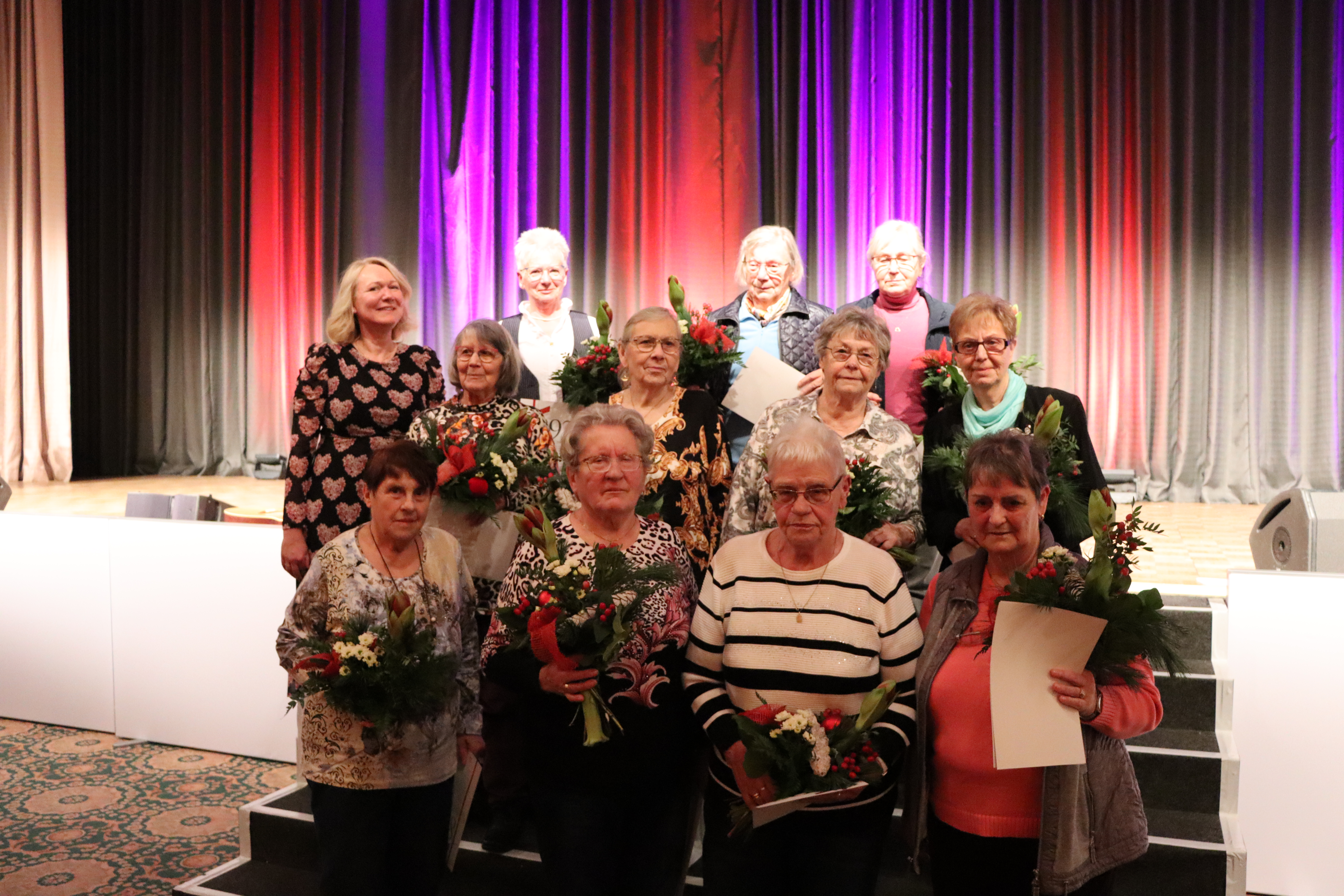 Zwölf ältere Frauen stehen auf einer Bühne mit bunten Vorhängen, halten Blumensträuße und Urkunden, posieren für ein Gruppenfoto.