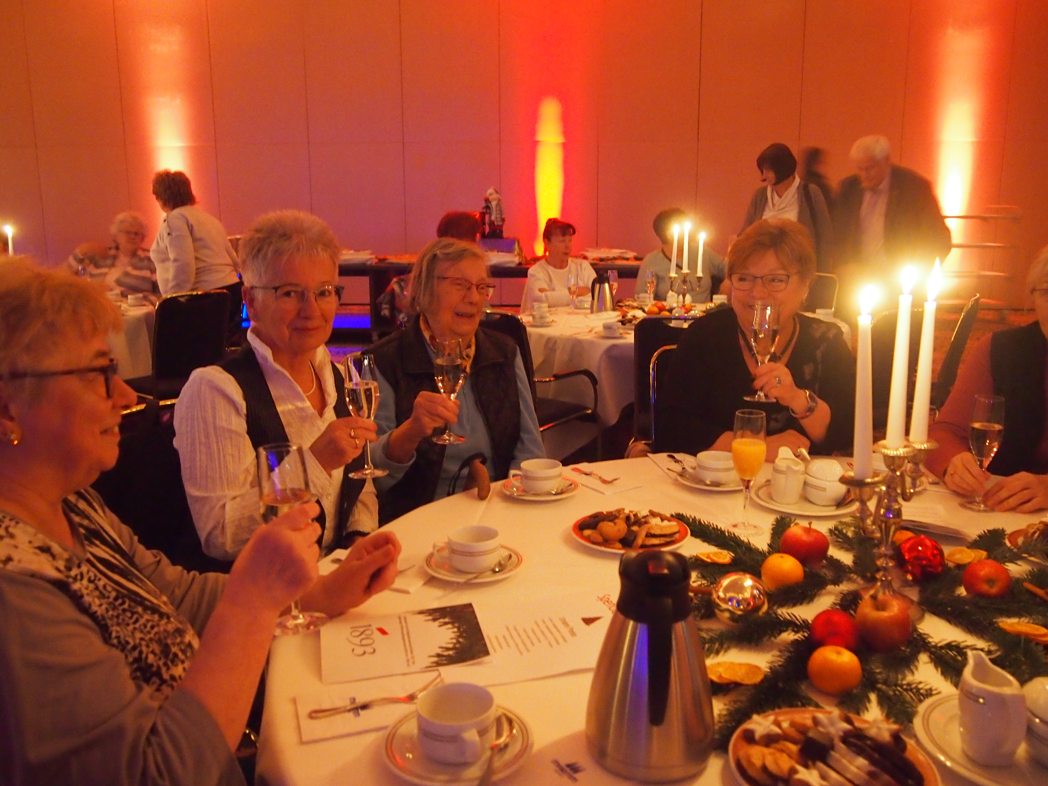 Ältere Frauen sitzen an einem festlich gedeckten Tisch mit Kerzen, trinken Sekt und unterhalten sich in einem warm beleuchteten Raum
