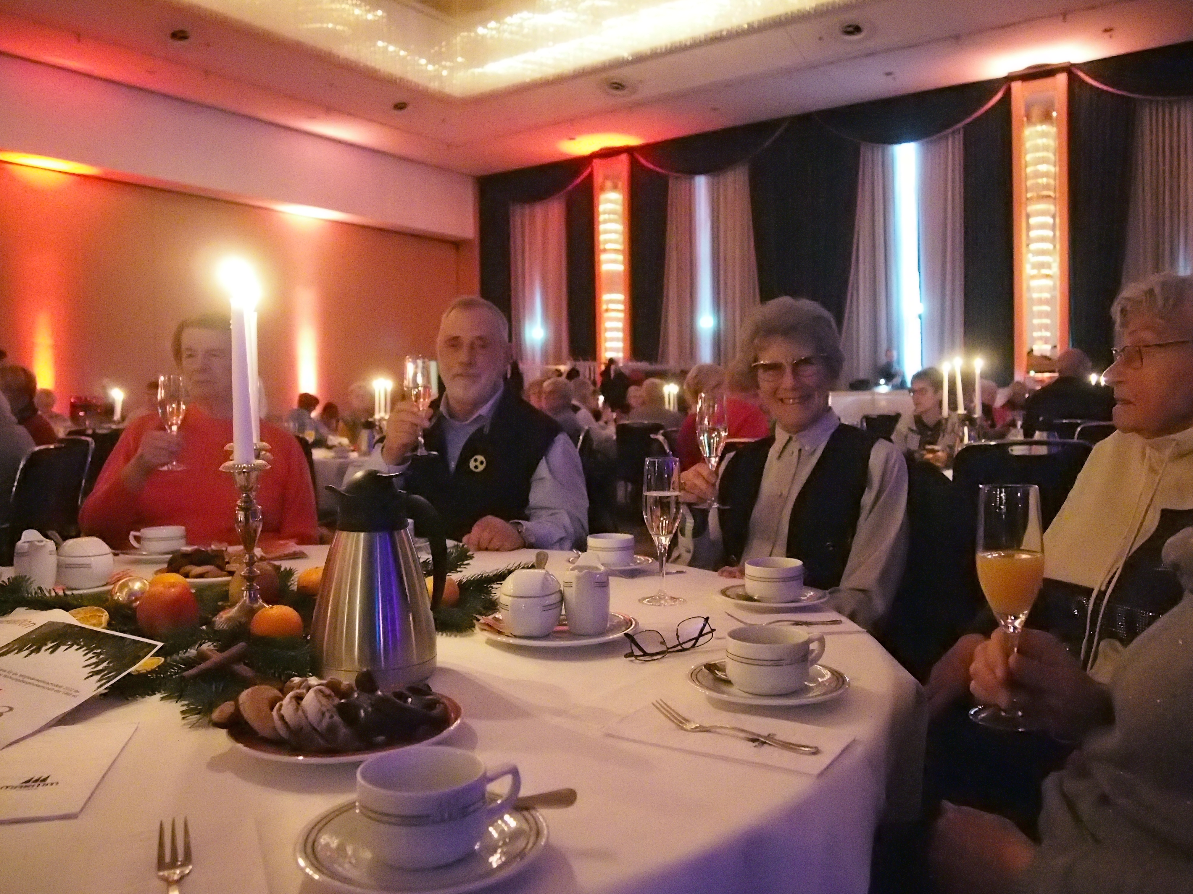 Ältere Menschen sitzen an einem festlich gedeckten Tisch mit Kerzen, Tassen und Gläsern, sie heben ihre Gläser zum Anstoßen in einem festlichen Saal