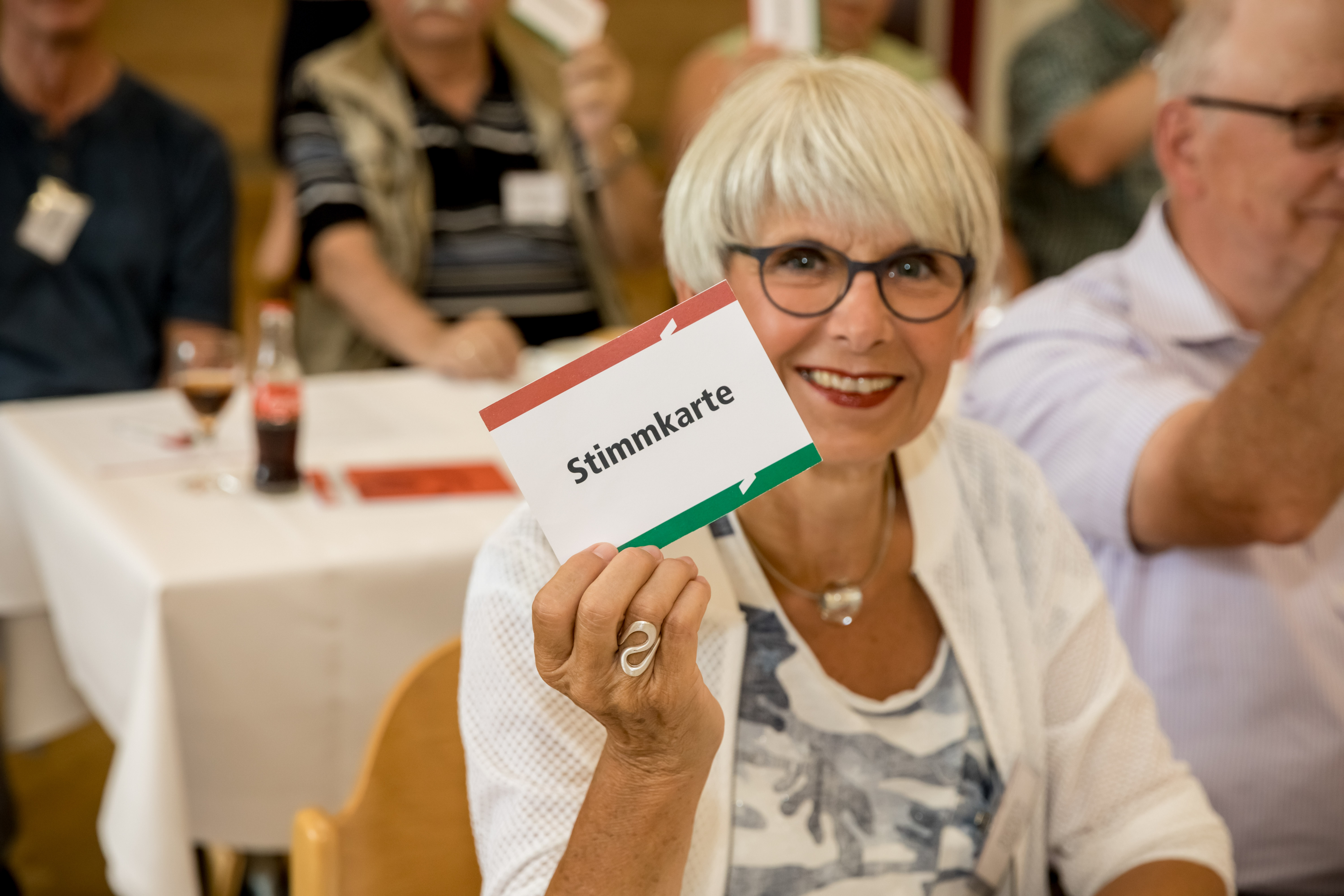 Ältere Frau mit Brille hält eine Stimmkarte hoch und lächelt in einem Versammlungsraum
