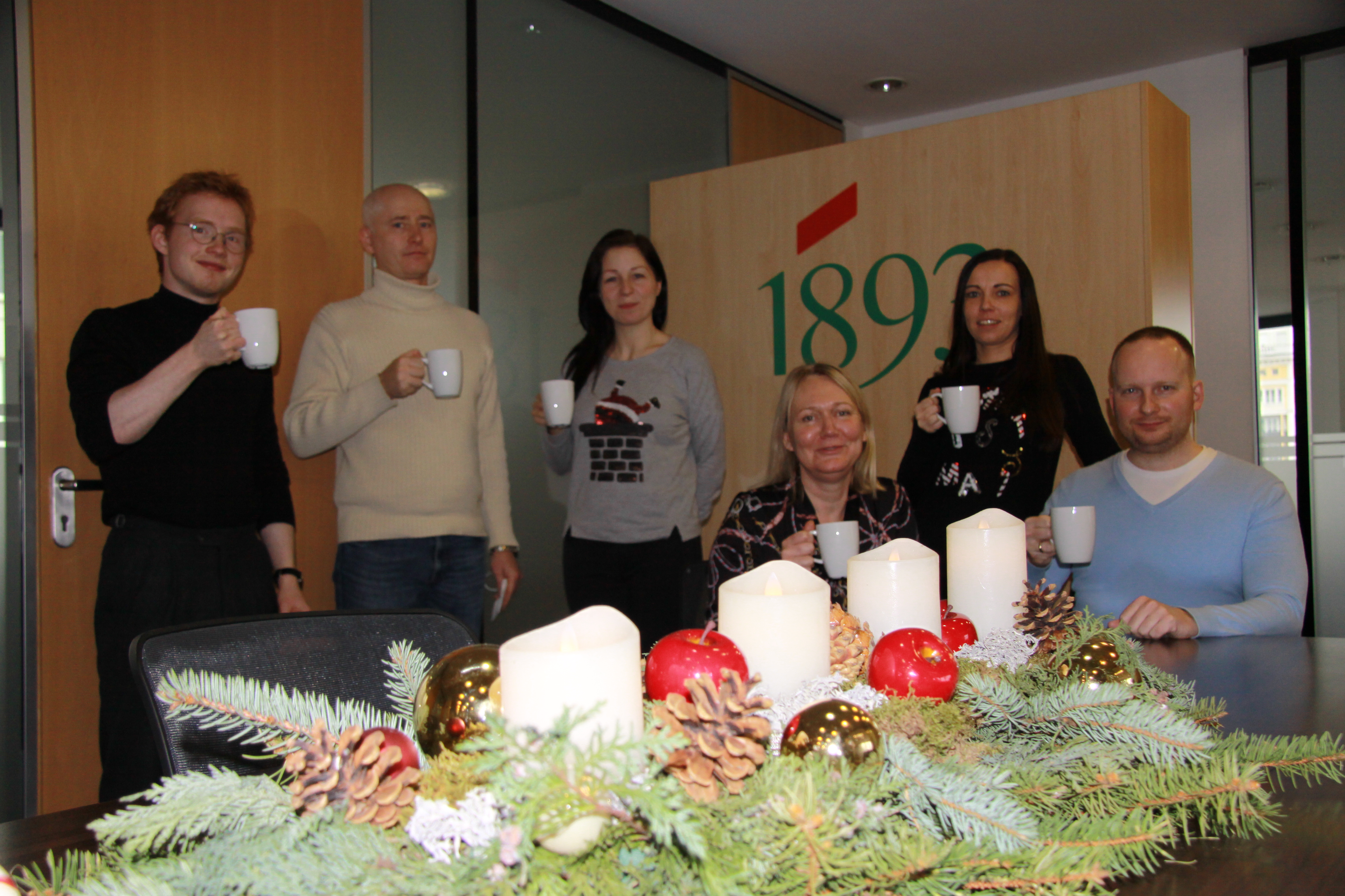Sechs Mitarbeiter der 1893 stehen und sitzen um einen Tisch mit Adventskranz und halten weiße Tassen, im Hintergrund eine Wand mit dem Logo der 1893 eG