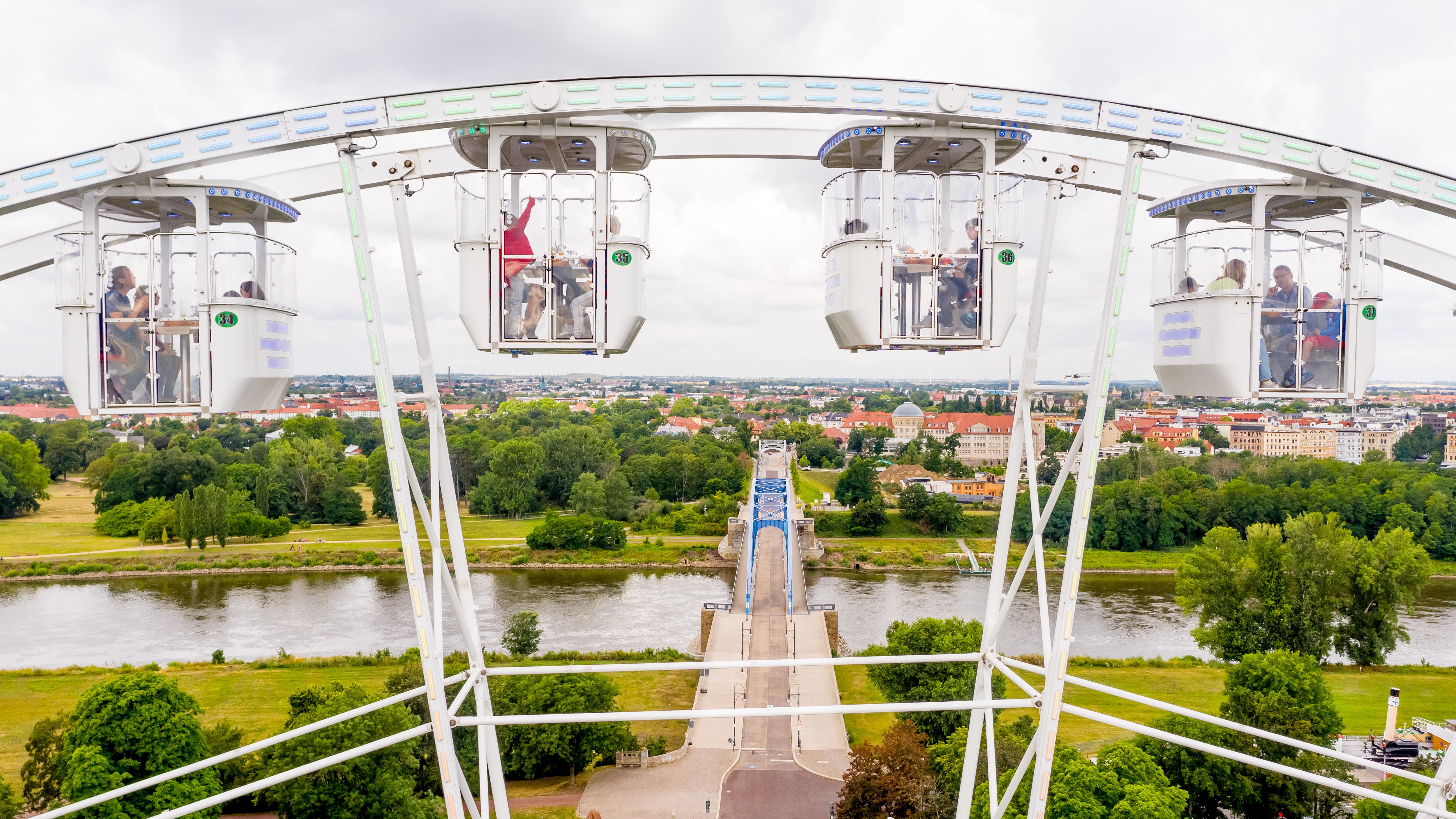 Weißes Riesenrad mit Gondeln in denen Fahrgäste sitzen vor einem Fluss und einer grünen Landschaft