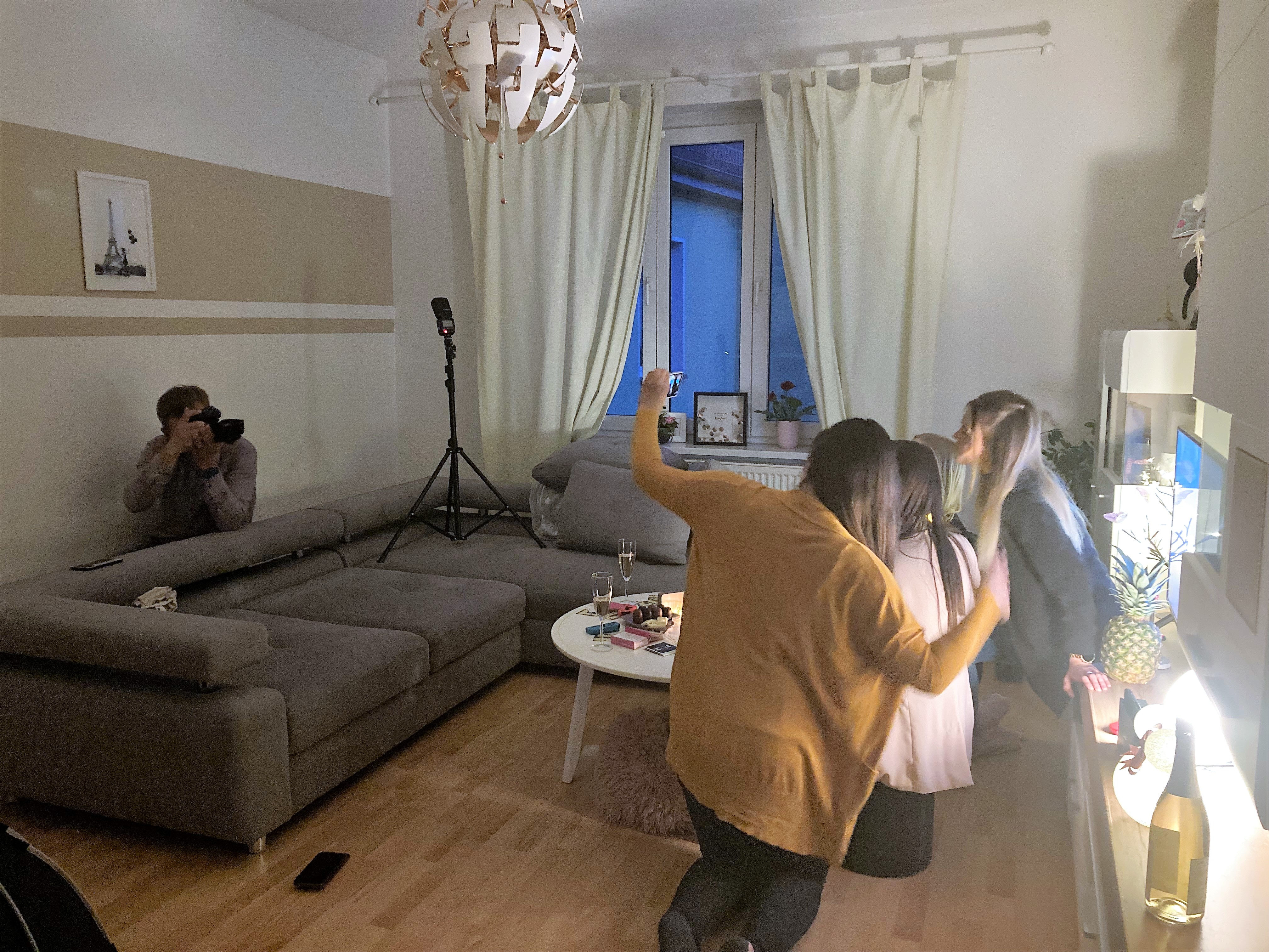 Drei Personen posieren in einem Wohnzimmer, während eine vierte Person mit der Kamera Fotos macht