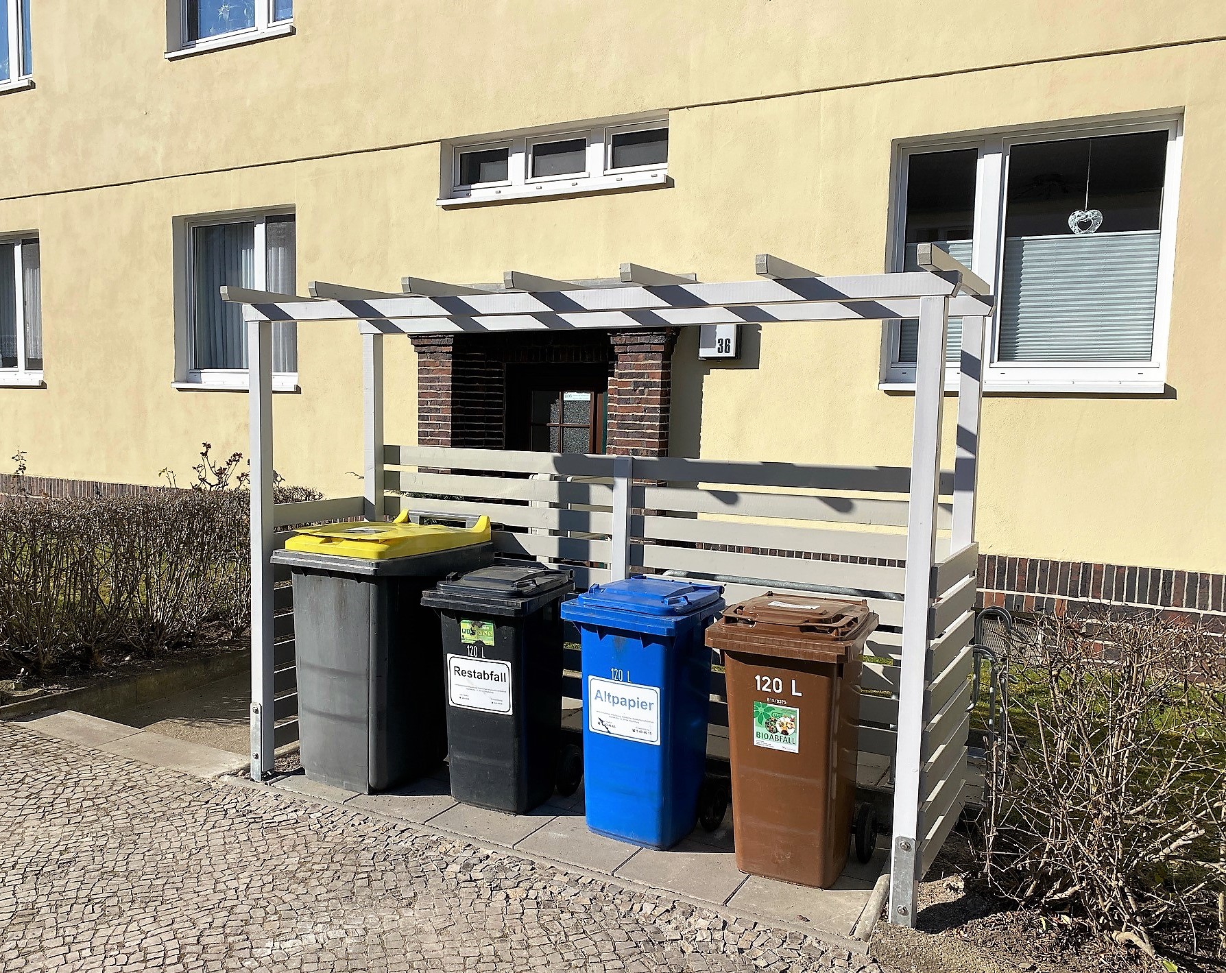 Vier Mülltonnen unter einem weißen Holzunterstand vor einem gelben Wohnhaus mit Fenstern, beschriftet für Restabfall, Altpapier und Bioabfall