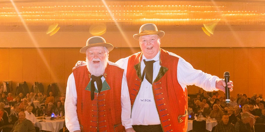 Zwei ältere Männer in traditionellen roten Westen und Hüten stehen auf einer Bühne, einer hält ein Mikrofon, im Hintergrund Publikum bei einer Weihnachtsfeier