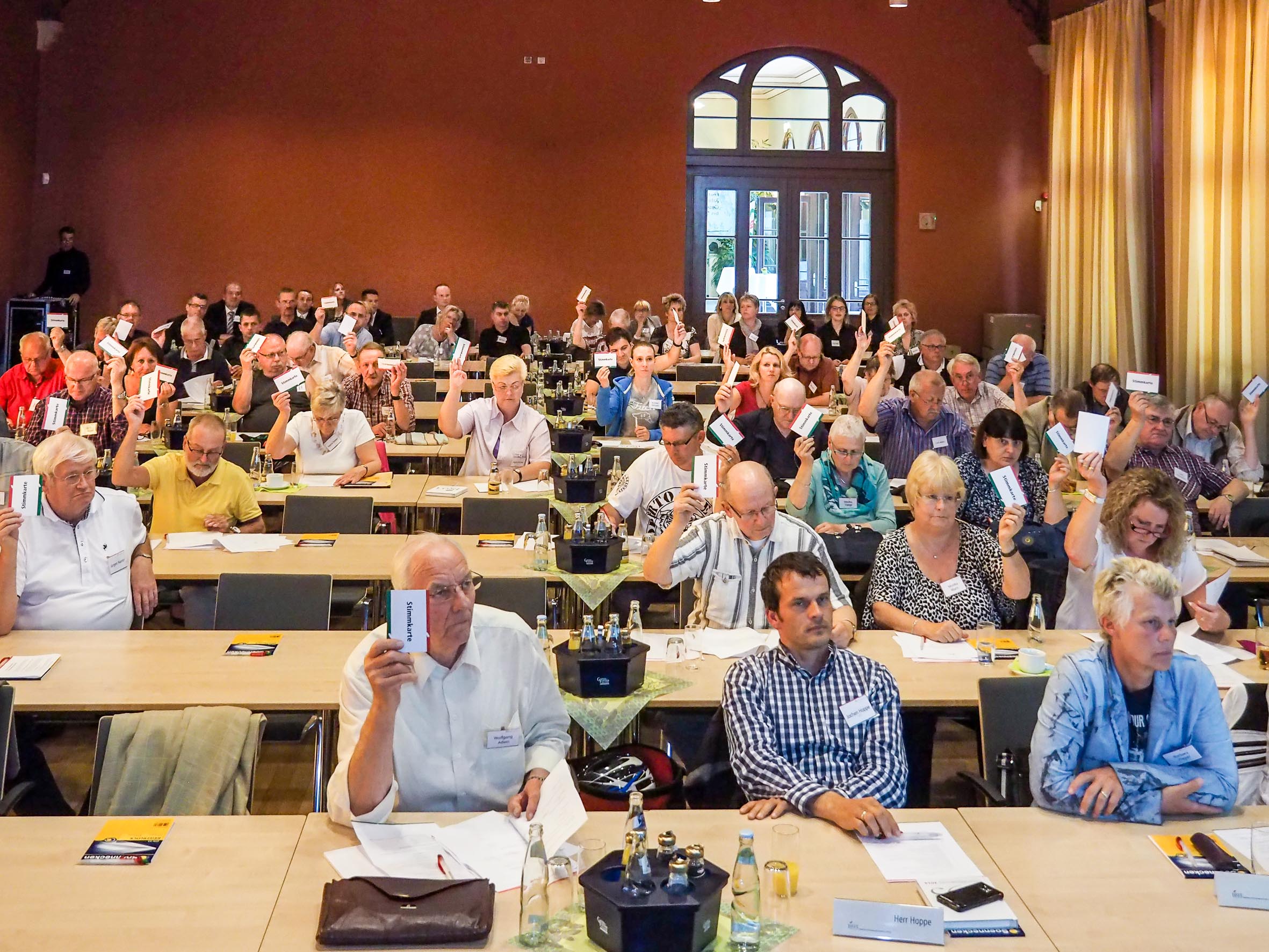 Menschen sitzen in Tischreihen in einem Saal und heben Stimmzettel in die Luft