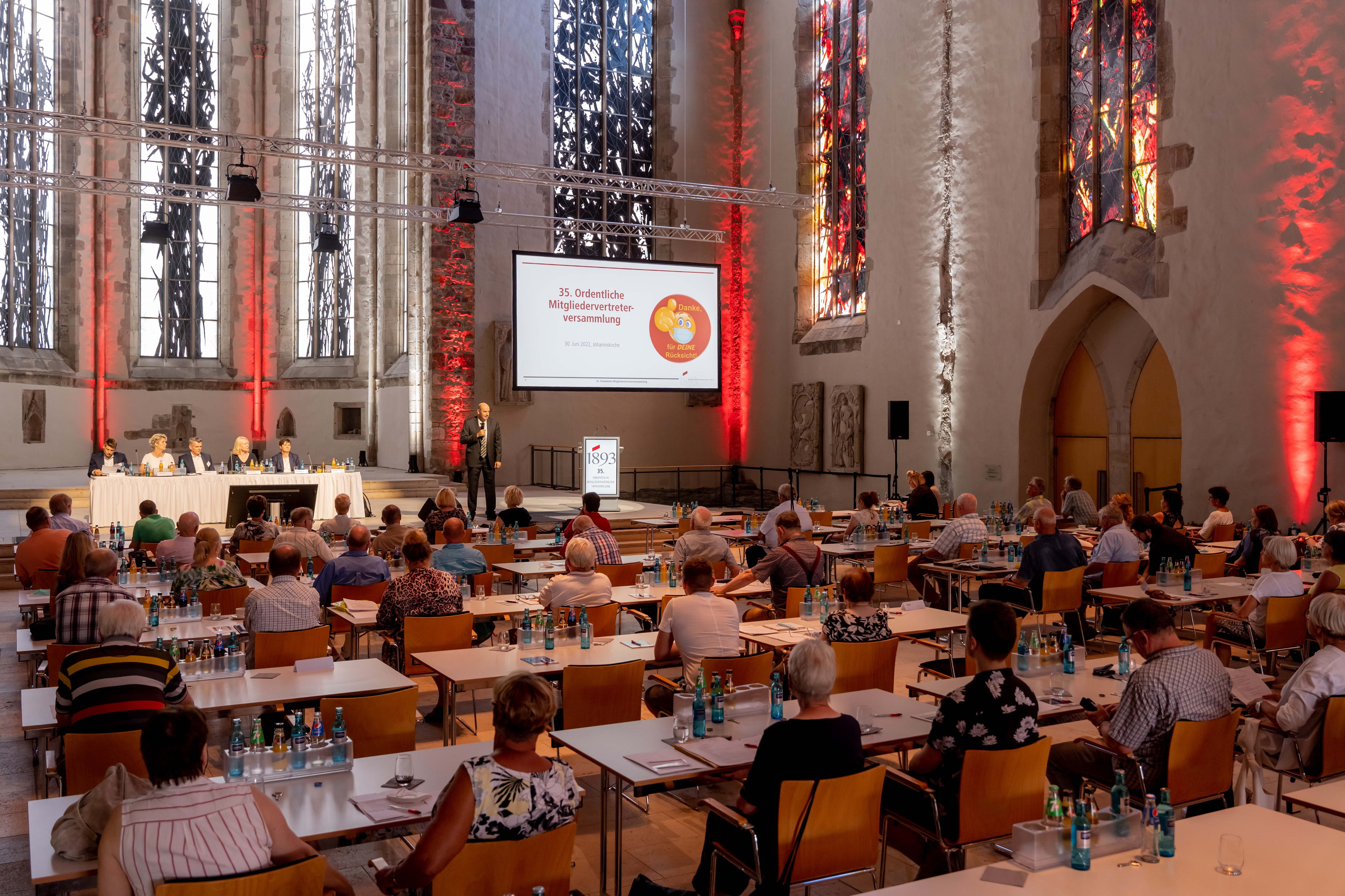 Menschen an Tischen in einem Saal mit hohen Fenstern und roter Beleuchtung, Präsentation auf Leinwand mit dem Titel '36. Ordentliche Mitgliederversammlung'
