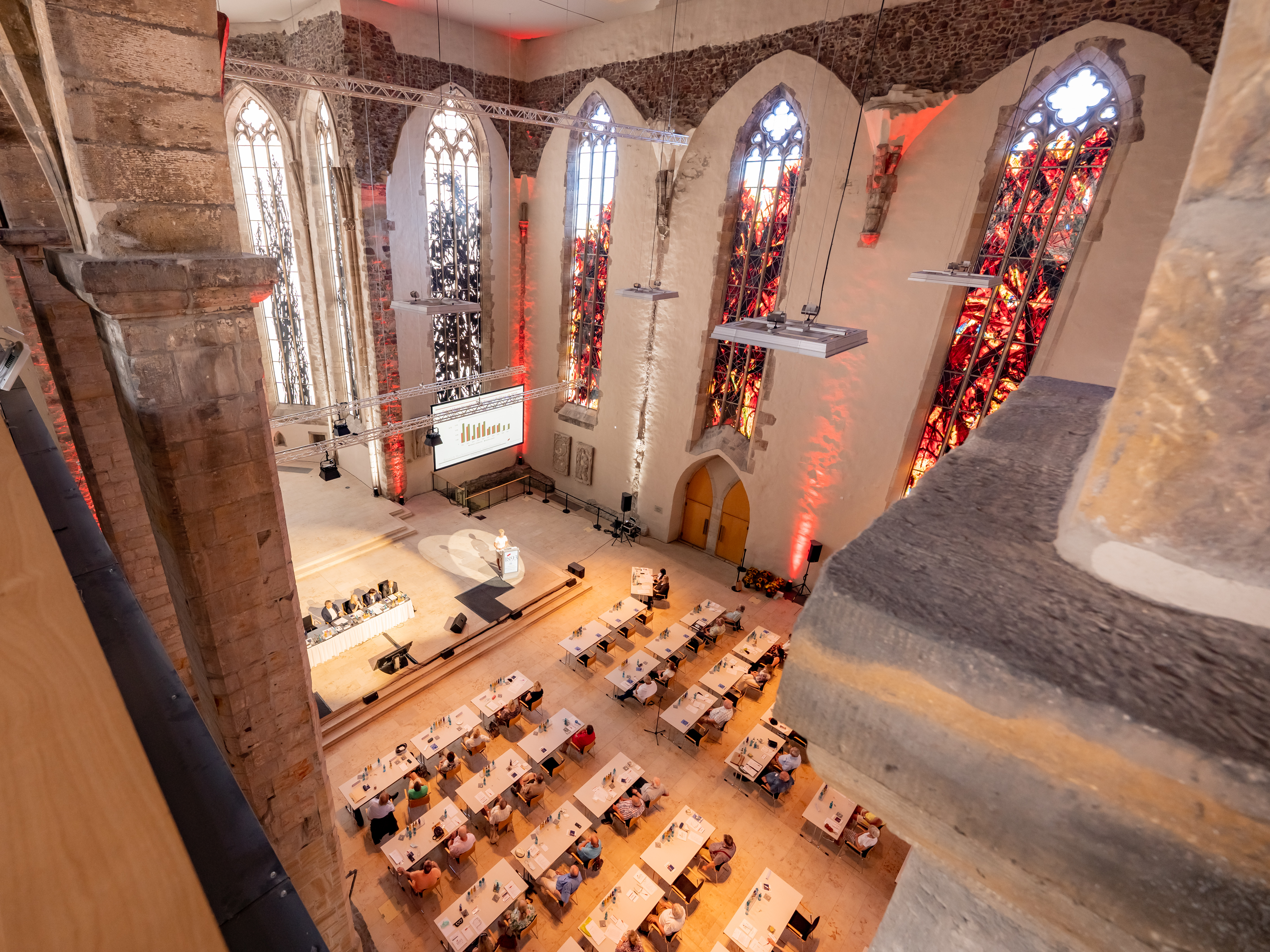 Blick von oben auf eine Veranstaltung in einer Kirche mit hohen gotischen Fenstern und mehreren Reihen von Tischen mit Teilnehmern