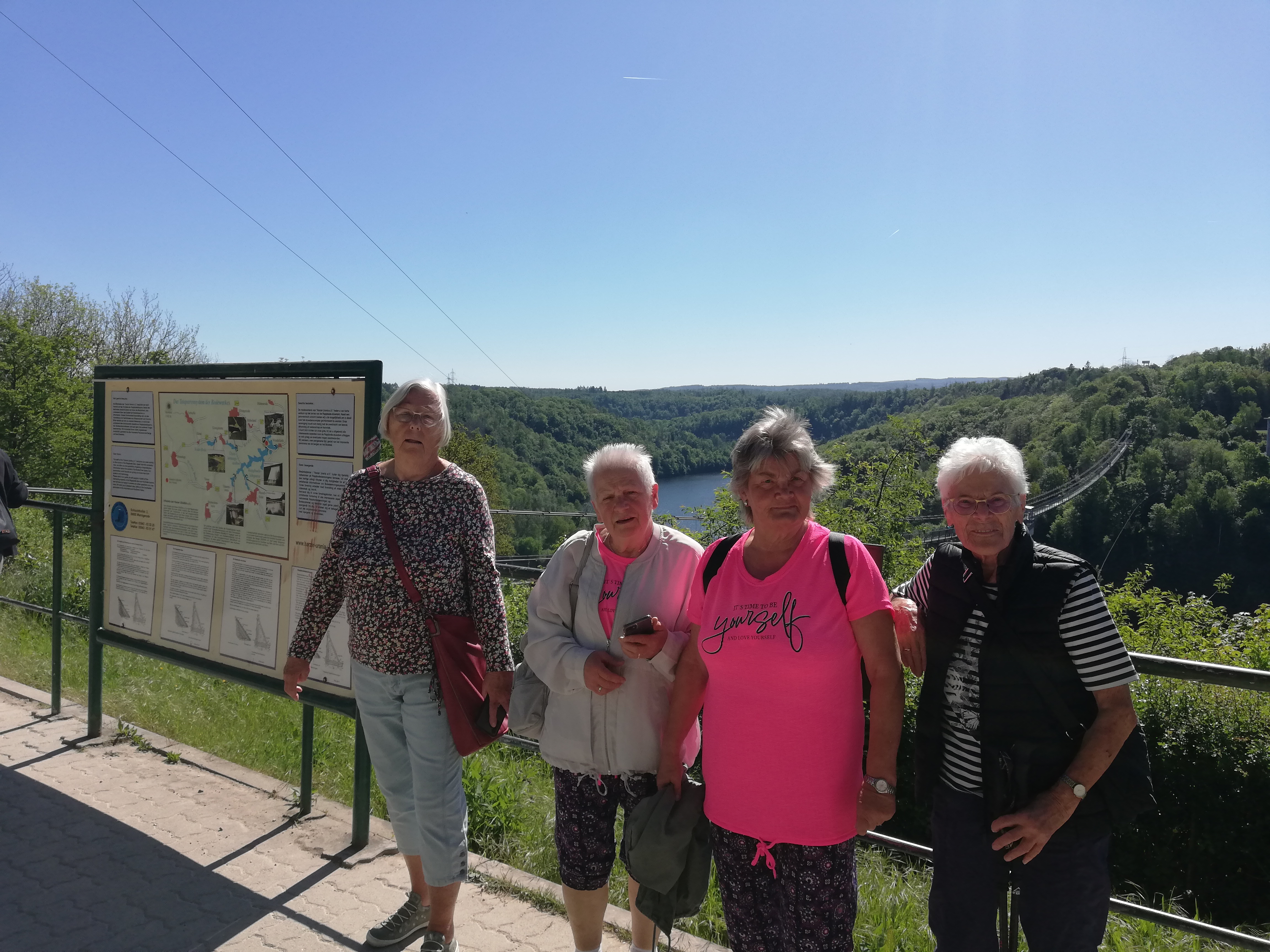 Vier ältere Frauen stehen vor der Rappbodetalsperre mit Wald und Hängebrücke im Hintergrund, zwei tragen pinke Oberteile.