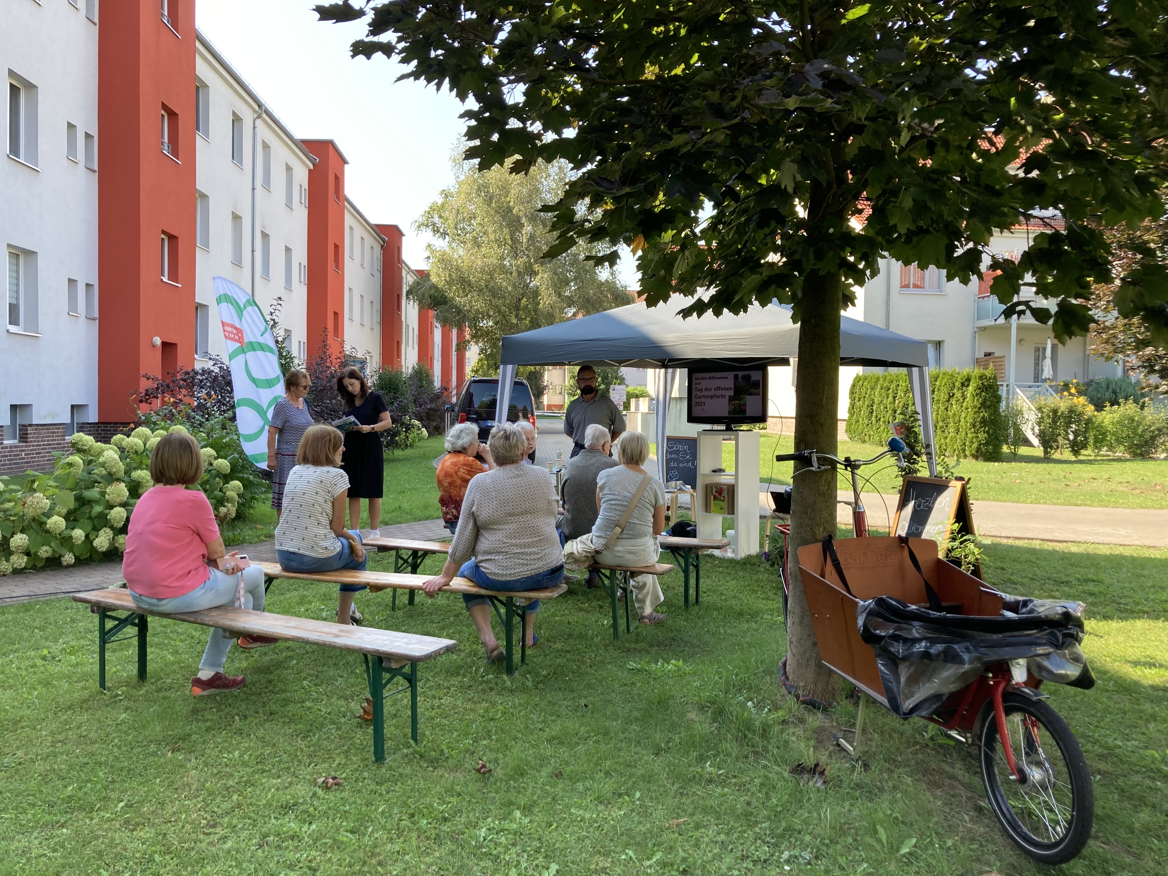 Mehrere Personen sitzen auf Bänken im Freien vor einem Pavillon mit einem Bildschirm, umgeben von Wohngebäuden und Bäumen