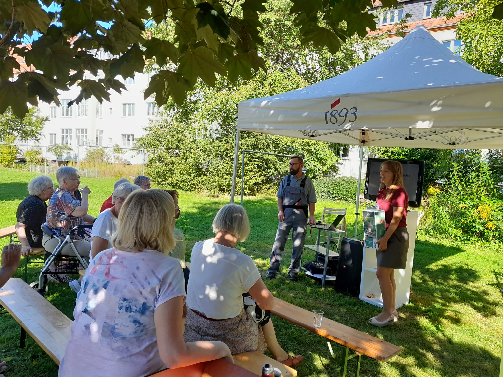 Seniorinnen sitzen auf Bänken im Freien und hören einer Frau zu, die unter einem weißen Zelt mit der Aufschrift '1893' spricht