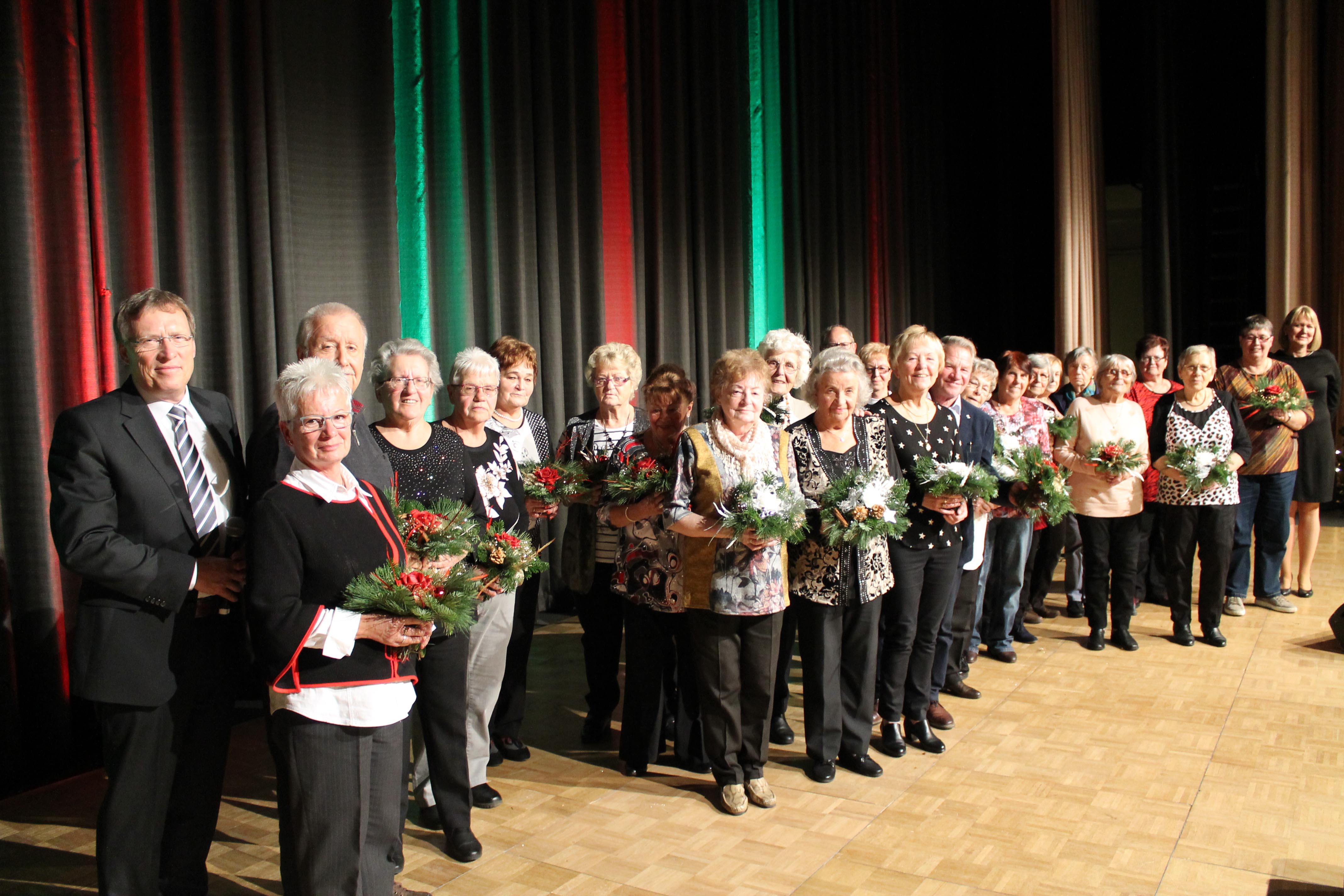 Eine Gruppe von älteren Menschen steht auf einer festlichen Bühne mit Blumensträußen in der Hand
