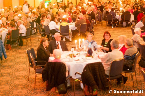 Menschen sitzen an einem festlich gedeckten Runden Tisch in einem Saal mit weiteren Menschen