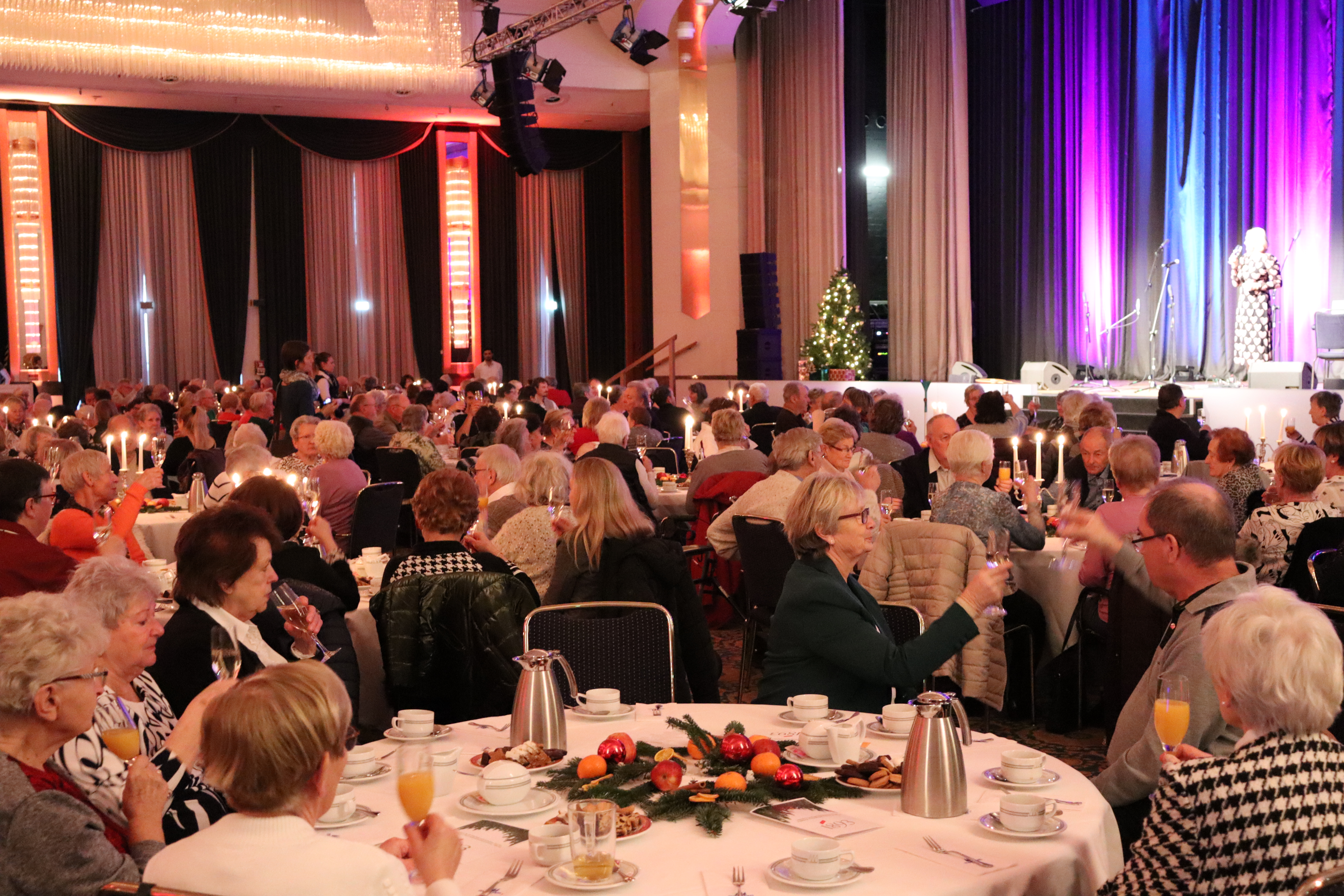 Menschen sitzen an festlich gedeckten Tischen in einem Saal mit Bühne und weihnachtlicher Dekoration
