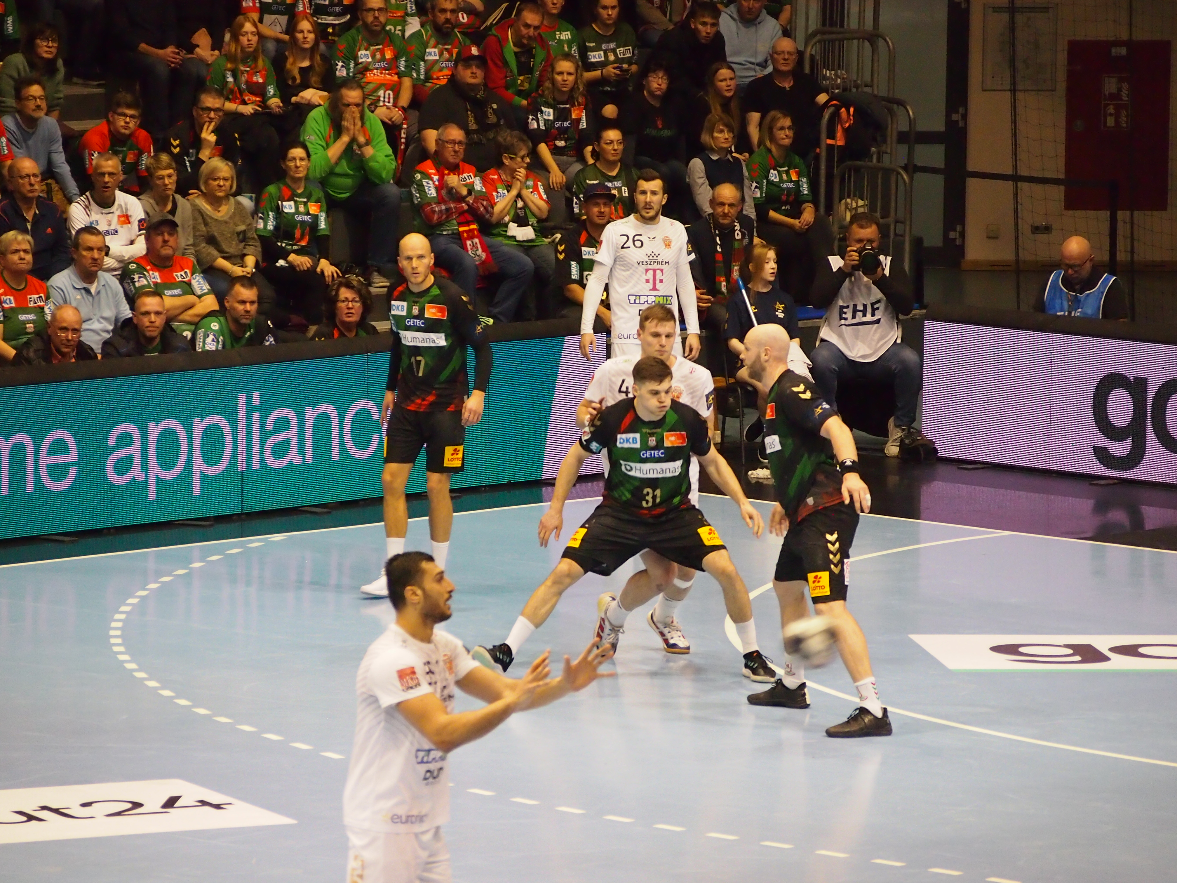 Handballspiel mit Spielern in weißen und schwarzen Trikots auf dem Spielfeld, Zuschauer im Hintergrund