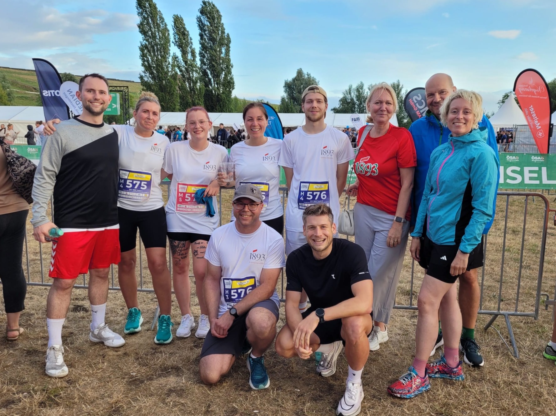 Neun Personen in Sportkleidung posieren auf einer Wiese bei einem Lauf-Event, einige tragen weiße T-Shirts mit '1893' und Startnummern