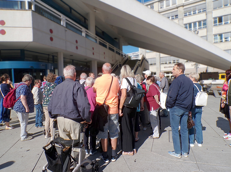 Eine größere Reisegruppe versammelt sich auf dem Platz am Berliner Fernsehturm. Die Teilnehmenden stehen im Freien nahe dem Eingangsbereich des Gebäudes und hören den Erläuterungen einer Führung zu. Einige Personen haben Taschen oder Rollatoren dabei. Im