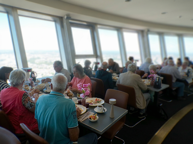 Mehrere Teilnehmende sitzen an gedeckten Tischen im Restaurant des Berliner Fernsehturms. Vor den großen Panoramafenstern genießen sie gemeinsam ihr Frühstück. Auf den Tischen stehen Kaffee, Brötchen, Aufschnitt, Obst und kleine Blumenvasen. Durch die Fen