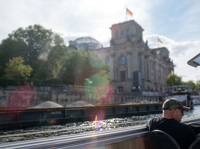 Blick von einem Ausflugsschiff auf die Spree im Berliner Regierungsviertel. Im Hintergrund steht ein historisches Gebäude mit deutscher Flagge auf dem Dach, dahinter ist die Glaskuppel des Reichstagsgebäudes zu erkennen. Im Vordergrund sind das Geländer d