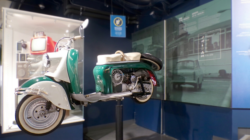 In einer Ausstellung des DDR Museums ist ein motorisierter Roller als Exponat zu sehen. Das Fahrzeug in Grün und Weiß steht erhöht auf einer Halterung und gibt durch eine geöffnete Seitenverkleidung den Blick auf den Motor frei. Im Hintergrund befinden si