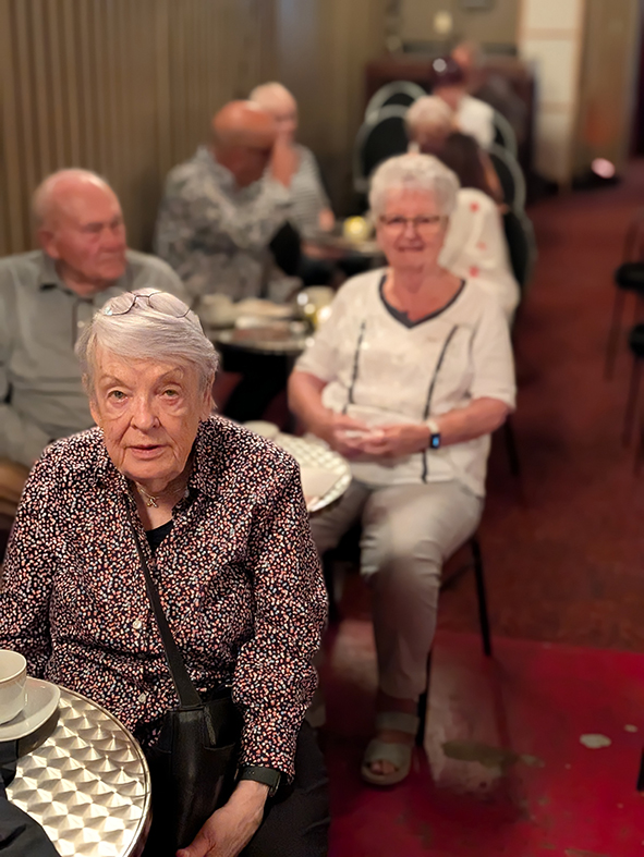 Mehrere Besucher in einem Kino- oder Veranstaltungssaal, die an kleinen Tischen hintereinander Platz genommen haben.