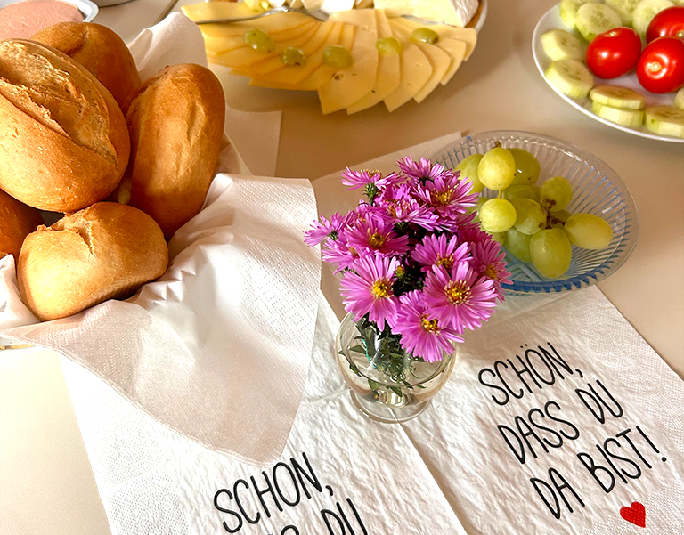 Brötchen, Obst, Käse und Blumen auf einer festlich gedeckten Frühstückstafel mit Serviettenaufdruck „Schön, dass du da bist!“