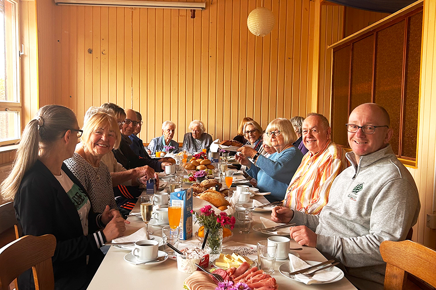 Teilnehmende eines Mieterfrühstücks sitzen an einer festlich gedeckten Tafel mit Brötchen, Eiern, Obst, Kaffee und Saftgetränken.