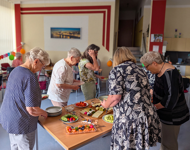Mehrere Teilnehmer bedienen sich an einem Buffet in einem Gemeinschaftsraum. Im Hintergrund sitzen weitere Gäste an Tischen und verbringen gemeinsam Zeit bei dem Mieterfrühstück.