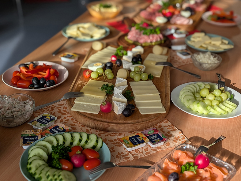 Mehrere Teilnehmer bedienen sich an einem Buffet mit verschiedenen Frühstücksspeisen. Auf dem Tisch stehen unter anderem Käse, Gemüse, Obst und weitere kleine Speisen.