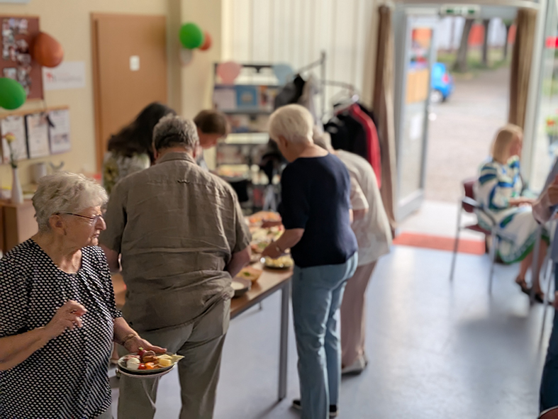 Mehrere Teilnehmer bedienen sich an einem Buffet in einem Gemeinschaftsraum. Im Hintergrund sind weitere Gäste sowie der Eingangsbereich des Raumes zu sehen.