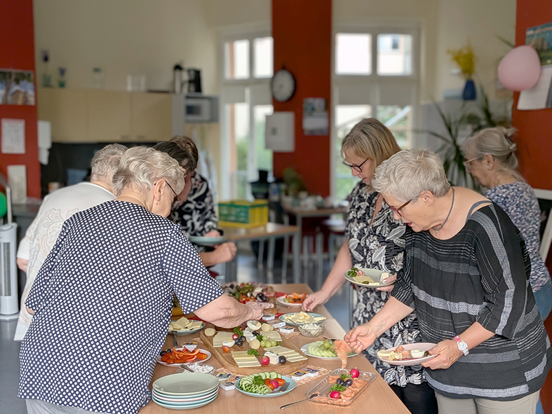 Mehrere Teilnehmer bedienen sich an einem Buffet mit verschiedenen Frühstücksspeisen. Auf dem Tisch stehen unter anderem Käse, Gemüse, Obst und weitere kleine Speisen.