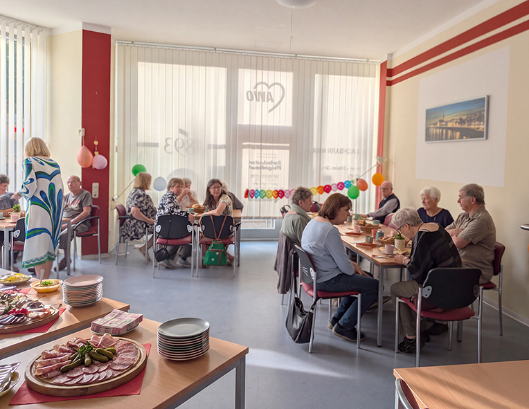 Mehrere Teilnehmer sitzen an gedeckten Tischen in einem Gemeinschaftsraum und frühstücken gemeinsam. Im Vordergrund ist ein Buffet mit verschiedenen Speisen zu sehen.