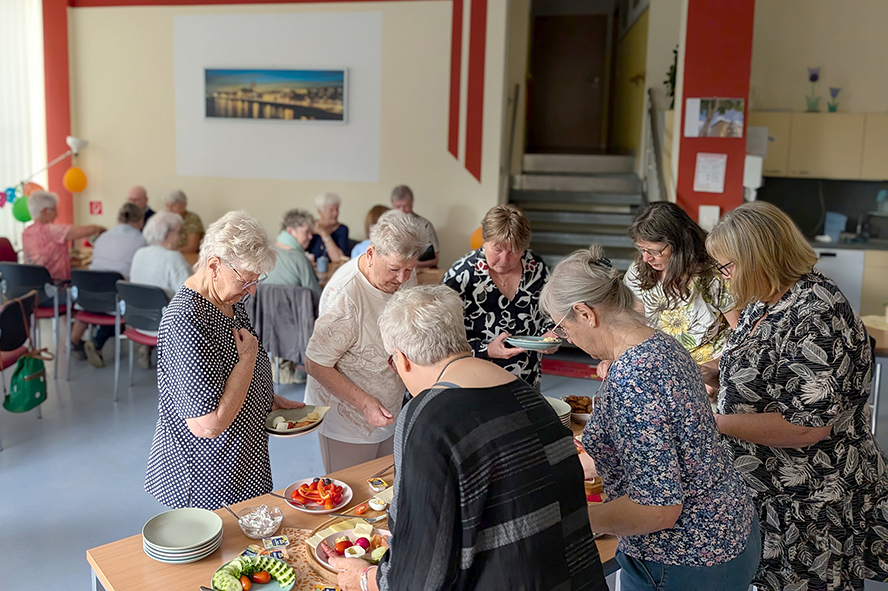 Mehrere Teilnehmer bedienen sich an einem Frühstücksbuffet in einem Gemeinschaftsraum. Im Hintergrund sitzen weitere Gäste an Tischen und verbringen gemeinsam Zeit bei der Veranstaltung.