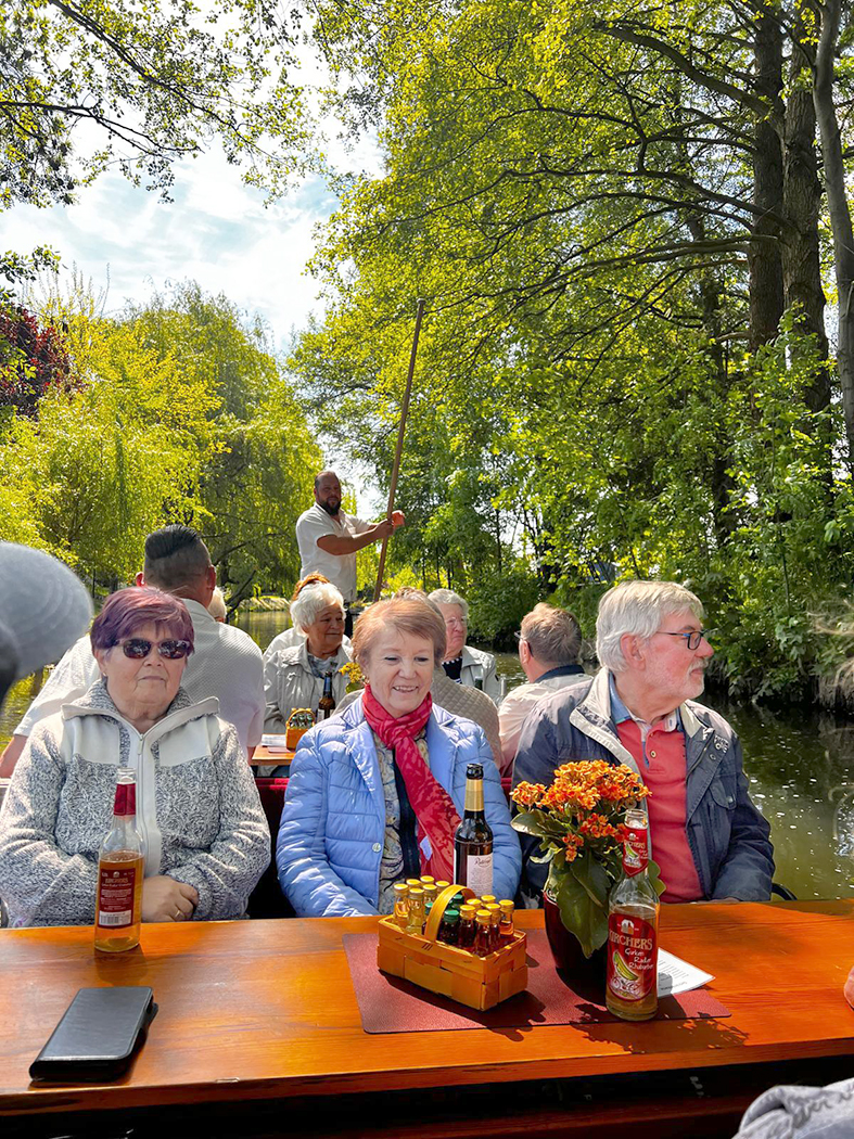 Teilnehmer einer Kahnfahrt sitzen an Tischen in einem überdachten Kahn und fahren durch einen schmalen, von Bäumen gesäumten Wasserlauf im Spreewald