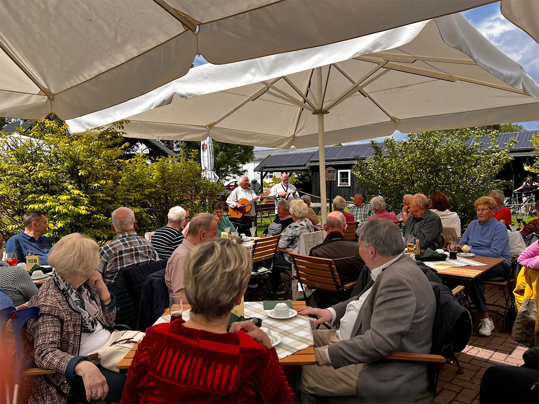Im Außenbereich eines Restaurants sitzen zahlreiche Gäste an Tischen unter Sonnenschirmen. Zwei Musiker sorgen mit Gitarrenmusik für Unterhaltung