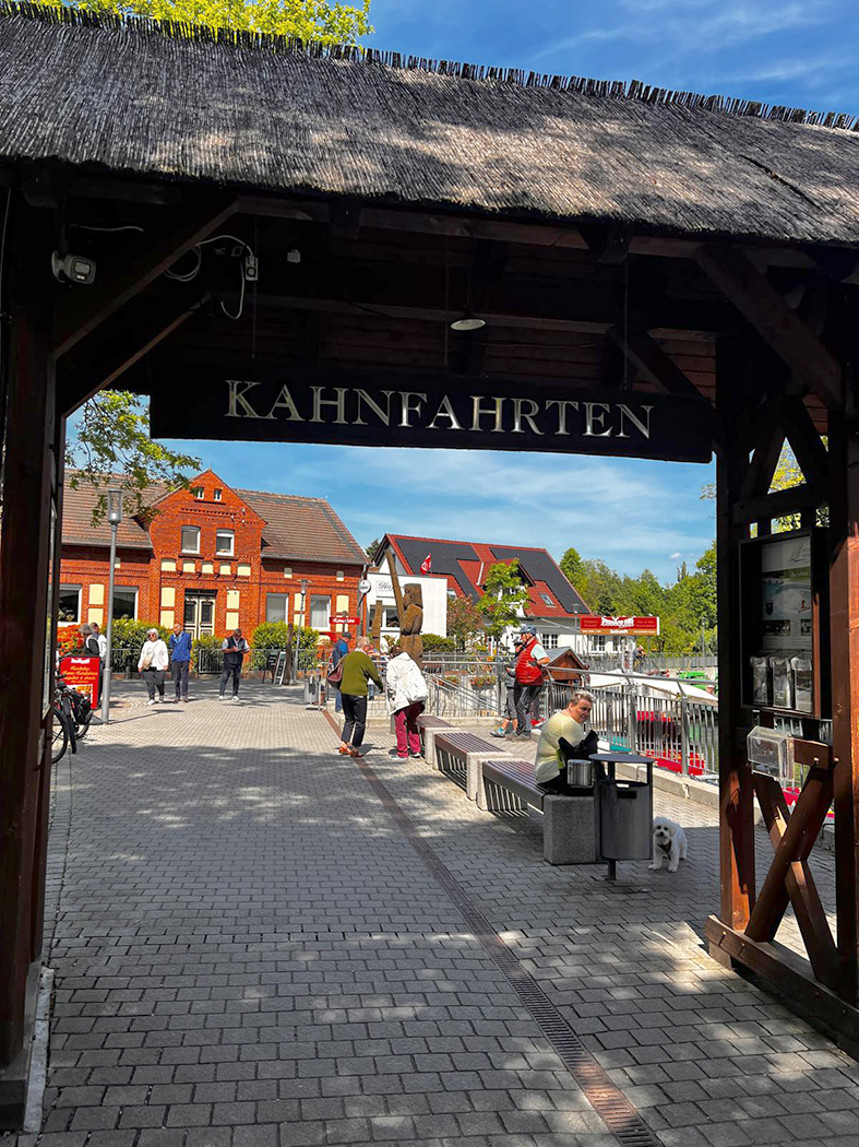 Im Vordergrund befindet sich ein großer überdachter Holzrahmen mit Reetdach. Unter dem Dach hängt ein schwarzes Schild mit der Aufschrift „KAHNFAHRTEN“, das den Weg zu den Anlegestellen weist.