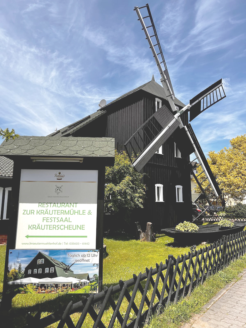 Eine historische Windmühle steht neben dem Restaurant Zur Kräutermühle. Vor dem Gebäude weist ein Schild auf Restaurant, Festsaal und Kräuterscheune hin