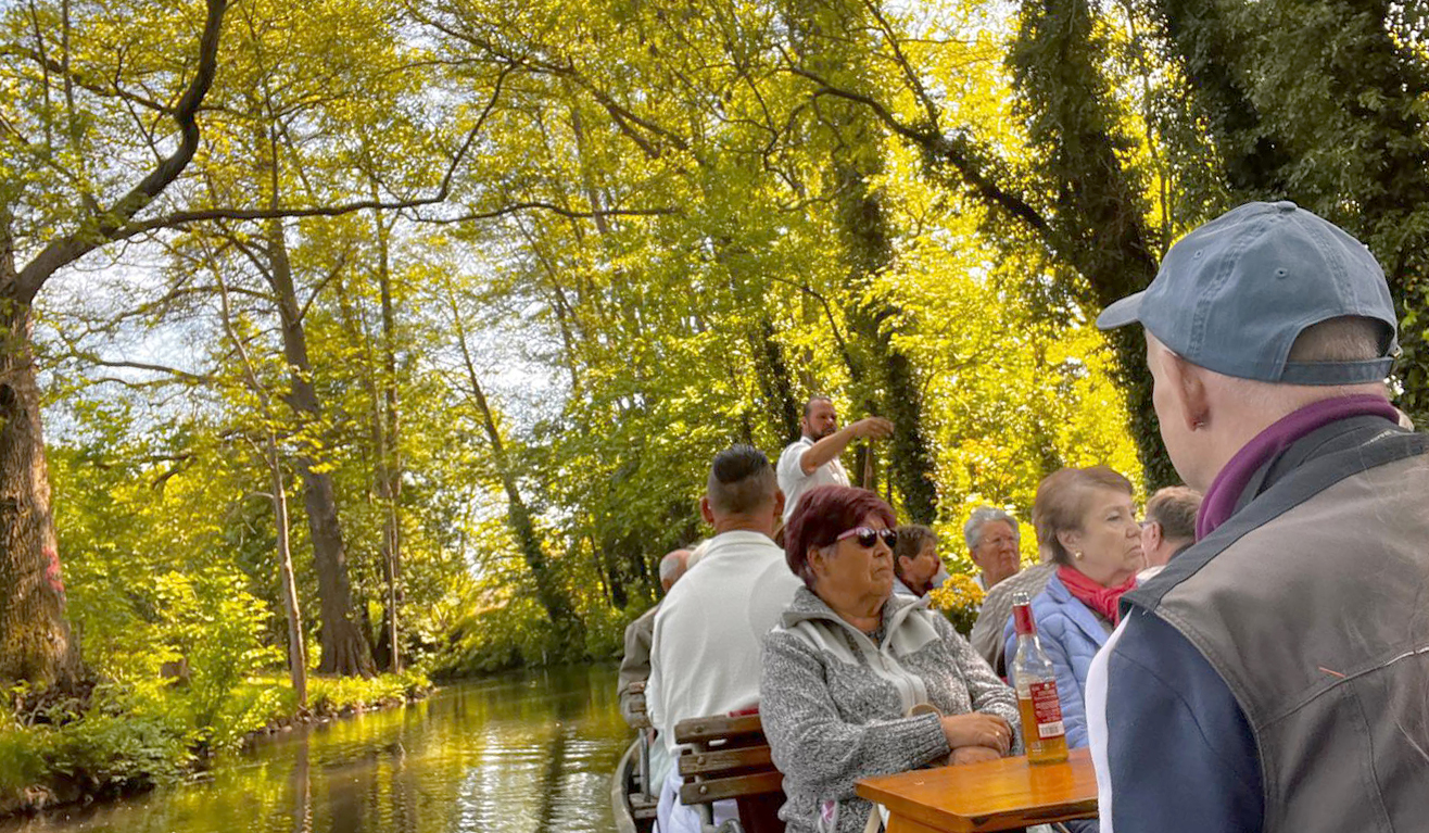 Gäste sitzen an Holztischen in einem traditionellen Kahn und genießen die Fahrt durch die von Bäumen und dichter Ufervegetation geprägte Landschaft. Im Hintergrund bewegt der Fährmann den Kahn mit einer langen Stange durch das ruhige Gewässer.