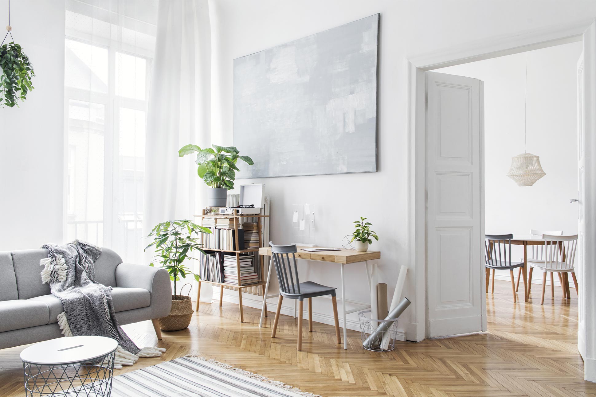 Helles Wohnzimmer mit grauem Sofa, Pflanzen, Schreibtisch mit Stuhl und großem abstraktem Bild an der Wand