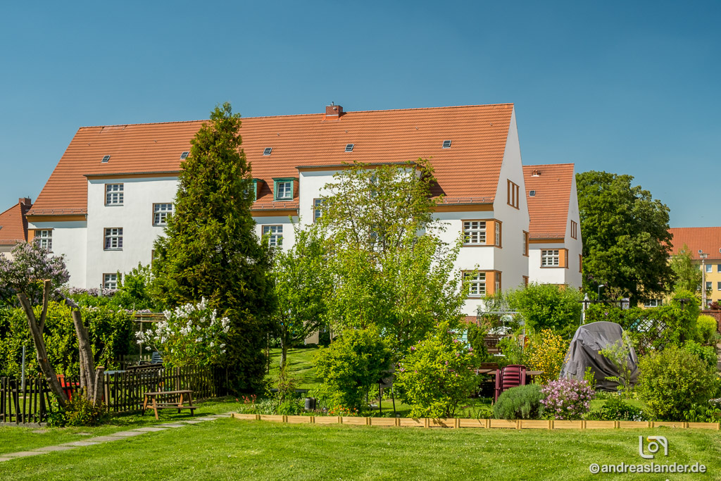 Weißes Wohngebäude mit rotem Ziegeldach hinter einem grünen Garten mit Bäumen und Sträuchern