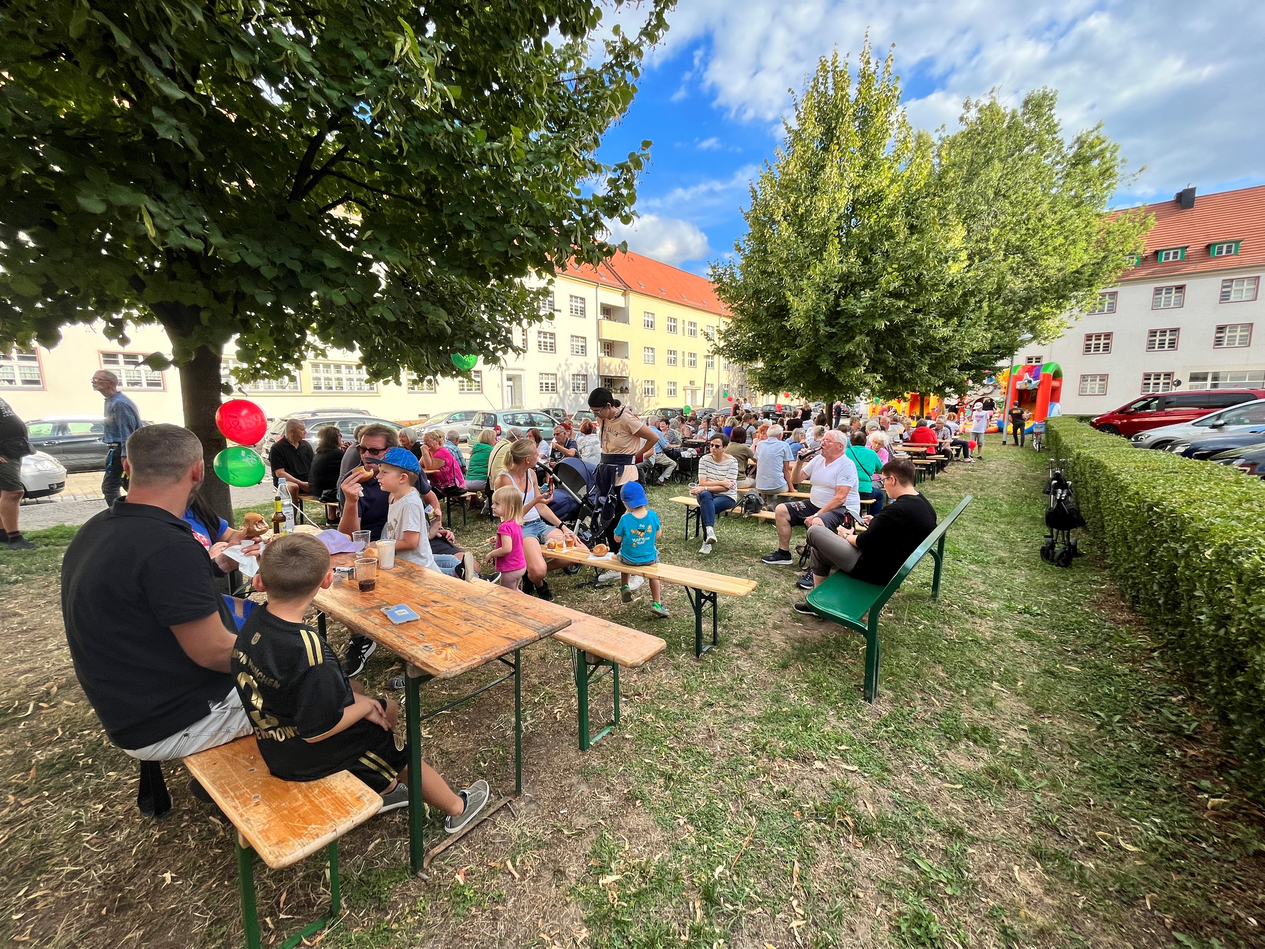 Menschen verschiedener Altersgruppen sitzen an Holzbänken im Freien bei einem Fest mit bunten Luftballons im Hintergrund