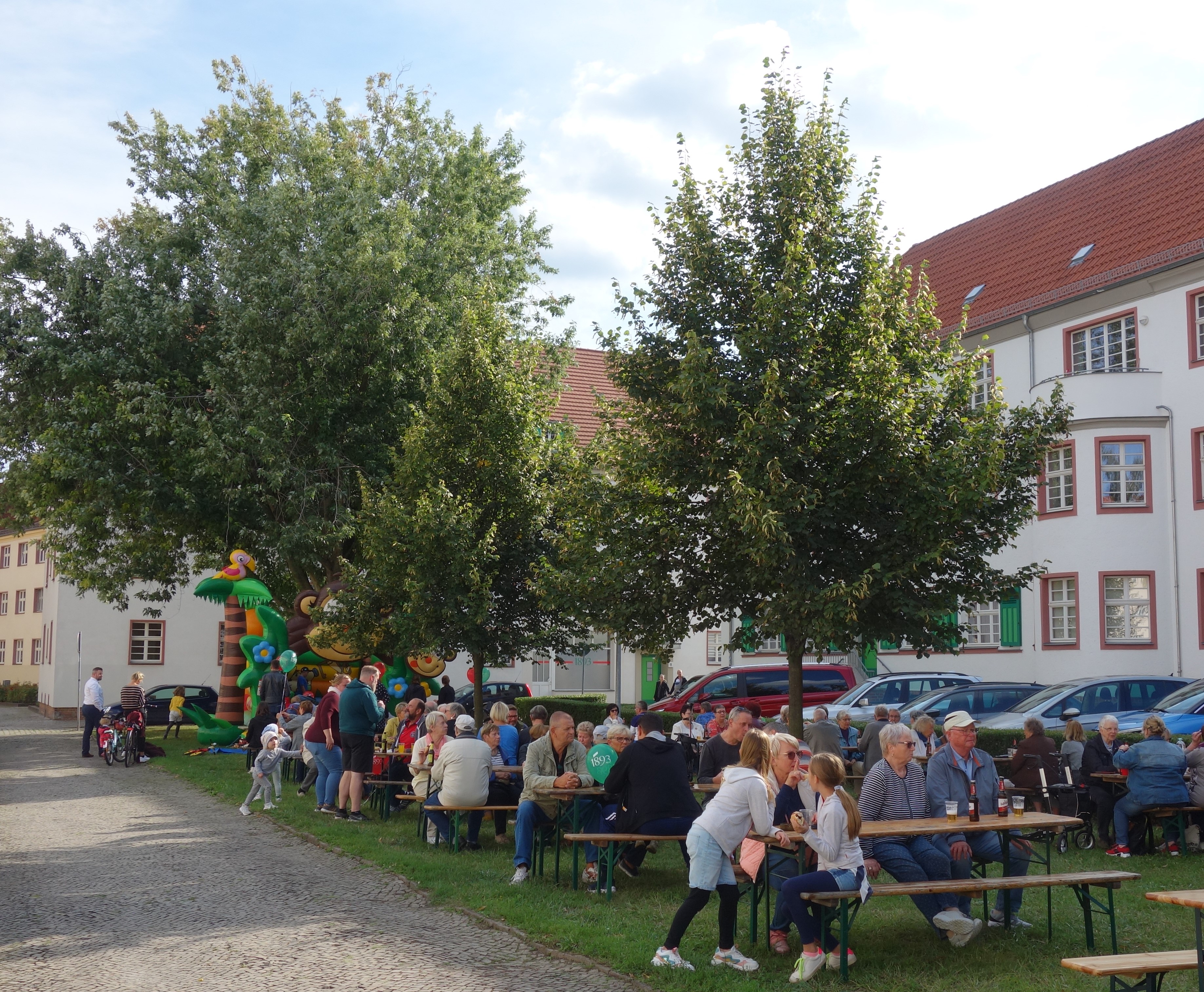 Mehrere Menschen sitzen an Bierbänken unter Bäumen. Im Hintergrund ist eine Hüpfburg und ein Wohngebäude.