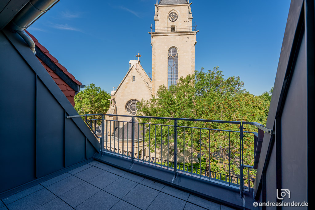 Blick von einem Balkon mit Metallgeländer auf eine Kirche mit Turmuhr und Bäumen bei klarem Himmel